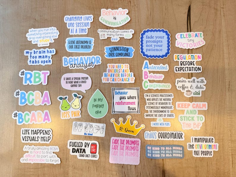 K&ouml;nnte beinhalten: Eine Sammlung bunter Aufkleber mit verschiedenen S&auml;tzen und Grafiken zum Thema Verhaltensanalyse. Die Aufkleber zeigen Texte wie "Behavior Analyst", "Celebrate Tiny Victories" und "Fueled by Data". Die Aufkleber sind auf einer hellbraunen Holzoberfl&auml;che angeordnet.