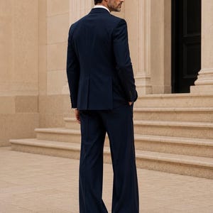 May include: A dark navy blue suit consisting of a tailored blazer and matching wide-leg trousers. The text at the bottom reads METRIC & MANOR Unlock Elegance. The suit features a structured back with a center vent and long sleeves.