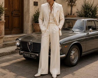 May include: A cream-colored suit consisting of a double-breasted blazer and wide-leg trousers, paired with a matching button-down shirt and cream shoes. The ensemble is worn by a person with dark hair and sunglasses standing beside a vintage gray car.