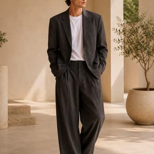 May include: A dark charcoal grey suit consisting of a single-breasted blazer with notched lapels and wide-leg trousers, worn over a plain white t-shirt. The outfit is paired with white trainers. The text METRIC & MANOR Unlock Elegance is visible.