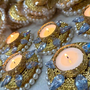 May include: Close-up of several lit candle holders. Each candle sits in a gold-colored filigree cup, surrounded by blue crystal accents and a ring of white pearls. The candles are lit, casting a warm glow.