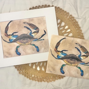 May include: Watercolor paintings of a blue crab, one framed in white and the other unframed. The crab has blue claws and legs, with a brown and tan body. The artwork is displayed on a woven, circular placemat.