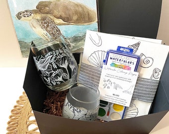 May include: A black gift box filled with a watercolor painting of a sea turtle, a set of watercolor paints, a paintbrush, two wine glasses with floral designs, and a coloring book with a sea turtle design. The box is sitting on a woven placemat.