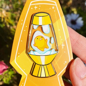 May include: A yellow lava lamp pin with a cartoon egg character inside. The lamp is outlined in white and has a gold base.