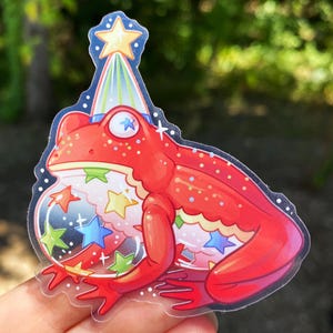 May include: A red frog sticker wearing a party hat with a star on top. The frog holds a bubble filled with colorful stars. The sticker has a clear border.