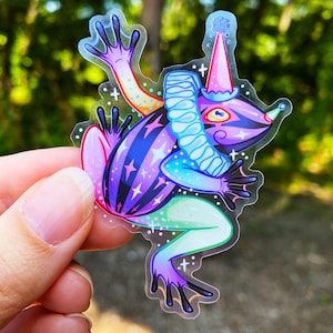May include: A colorful sticker featuring a whimsical frog character. The frog is adorned with a party hat, a ruffled collar, and a star-patterned body. The sticker has a transparent background and is held by a hand.