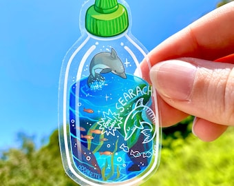 May include: A clear plastic bottle sticker with a blue and green ocean scene. The sticker features a dolphin, a shark, seaweed, and fish. The text "Real Cool!" and "Searach" are on the sticker.