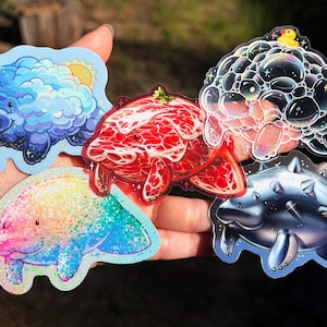 May include: A collection of five manatee-shaped stickers featuring diverse designs: a blue cloud manatee with a sun, a red marbled meat-textured manatee, a bubble-covered manatee with a yellow duck, a rainbow glitter manatee, and a metallic spiked manatee.