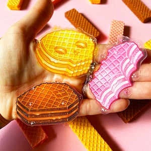May include: Three waffle cookie stickers with a glittery finish. The stickers are shaped like a skull, a stack of waffles, and a stack of pink waffles. The stickers are on a pink surface.