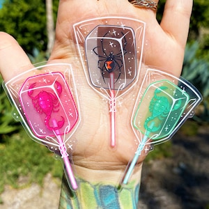 Puede incluir: Tres pegatinas de plástico transparente con forma de piruleta con fondos de colores diferentes y diseños de insectos. La primera pegatina tiene un fondo negro con una araña viuda negra. La segunda pegatina tiene un fondo rosa con un escorpión. La tercera pegatina tiene un fondo verde con un escorpión.