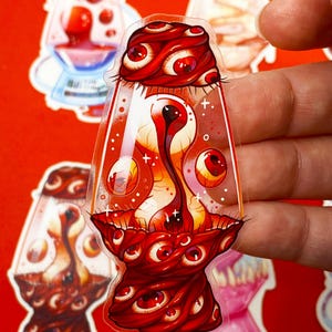 Pu&ograve; includere: Un adesivo trasparente con un design stilizzato di una lampada a lava nei toni del rosso e dell'arancione. La lampada &egrave; decorata con numerosi occhi rossi e una struttura centrale a forma di occhio. Altri adesivi sono visibili sullo sfondo.