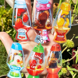 Könnte beinhalten: Sechs klare Glas-Lavalampe mit verschiedenen farbigen Flüssigkeiten und Designs. Die Designs beinhalten ein Schwein mit Kochmütze, eine Ketchupflasche, eine Senfflasche, eine Ranch-Dressing-Flasche, eine Sriracha-Flasche und eine Sojasauce-Flasche.