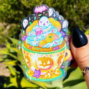 May include: A clear sticker featuring a Halloween-themed design. The sticker depicts a cup filled with a blue and orange concoction, topped with pumpkins, ghosts, and candy. The cup has a jack-o'-lantern design and a purple cat.