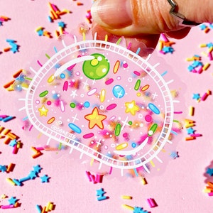 Puede incluir: Una pegatina transparente con fondo rosa y contorno blanco. La pegatina presenta un círculo lleno de chispas de colores, estrellas y otras formas. El texto "ODDARETTE" es visible en la parte inferior de la pegatina.