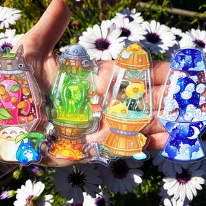 May include: Four colorful glass-like stickers with a whimsical design. Each sticker features a different scene inside a clear glass-like container. The first sticker features a pink and green Totoro character, the second sticker features a green alien-like creature, the third sticker features a yellow duck in a blue container, and the fourth sticker features a blue and white ghost-like creature.
