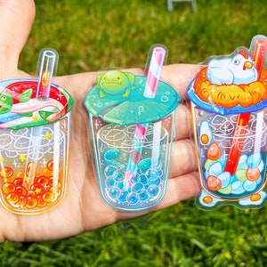 May include: Three clear plastic stickers of boba tea cups with different toppings. The first cup has a green fish and orange boba pearls. The second cup has a green frog and blue boba pearls. The third cup has a white chicken and yellow and blue boba pearls.