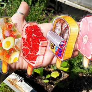 May include: Four clear plastic stickers with different food items on them. The stickers are shaped like popsicles and feature a clear jelly salad, a steak, a can of sardines, and a ham.
