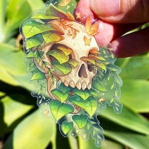 May include: A clear sticker with a white skull surrounded by green leaves and small orange butterflies. The skull is partially obscured by the leaves.