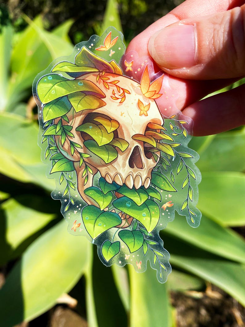 May include: A clear sticker with a white skull surrounded by green leaves and small orange butterflies. The skull is partially obscured by the leaves.