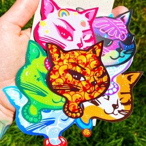 Può includere: Cinque adesivi di gatto colorati con diversi motivi e design. Gli adesivi presentano un gatto rosa con un arcobaleno e stelle, un gatto verde con stelle, un gatto rosso e arancione con un motivo a maccheroni, un gatto viola con una foglia tropicale e un gatto giallo con un campanello.