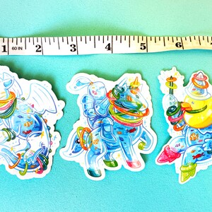 May include: Three colorful stickers featuring a blue dragon, a knight riding a unicorn, and a yellow creature with a crown. All three figures are surrounded by colorful rings and have a blue, watery background with fish.
