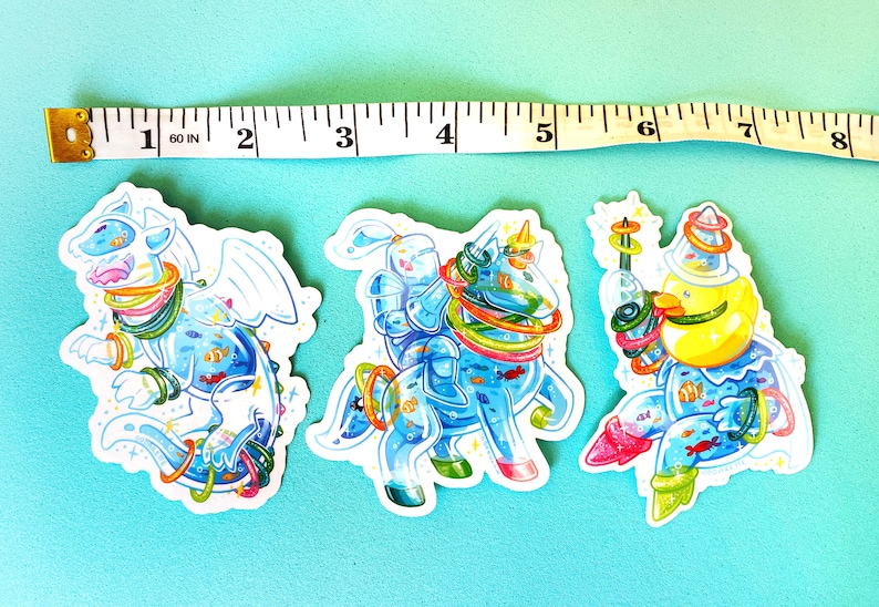 May include: Three colorful stickers featuring a blue dragon, a knight riding a unicorn, and a yellow creature with a crown. All three figures are surrounded by colorful rings and have a blue, watery background with fish.