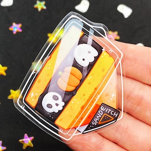 May include: A clear sticker featuring a Halloween-themed sandwich design. The sandwich has orange bread, a dark filling, and is decorated with a pumpkin and two skulls. The sticker has the text "SANDWITCH" on a triangular label.