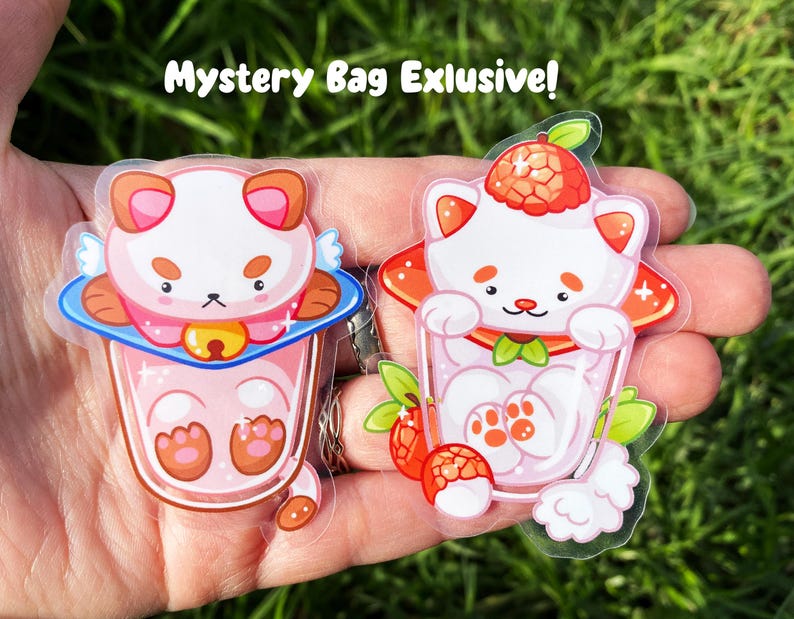 May include: Two clear stickers featuring cartoon illustrations of adorable white cats with pink accents. The cats are depicted in a glass of pink milk with a strawberry on top. The stickers are labeled "Mystery Bag Exclusive!"