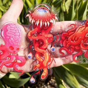 Peut inclure: Trois autocollants translucides colorés représentant un poulpe rose ressemblant à un cerveau, une créature rouge tentaculaire avec un œil et un poulpe rouge avec une longue tentacule flottante. Les autocollants sont sur une main.