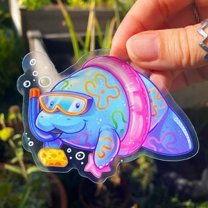 May include: A clear sticker featuring a cartoon manatee wearing a pink inflatable ring and goggles. The manatee is blue with a yellow sponge and pink starfish. The sticker has the text "Oddskate" in the bottom right corner.