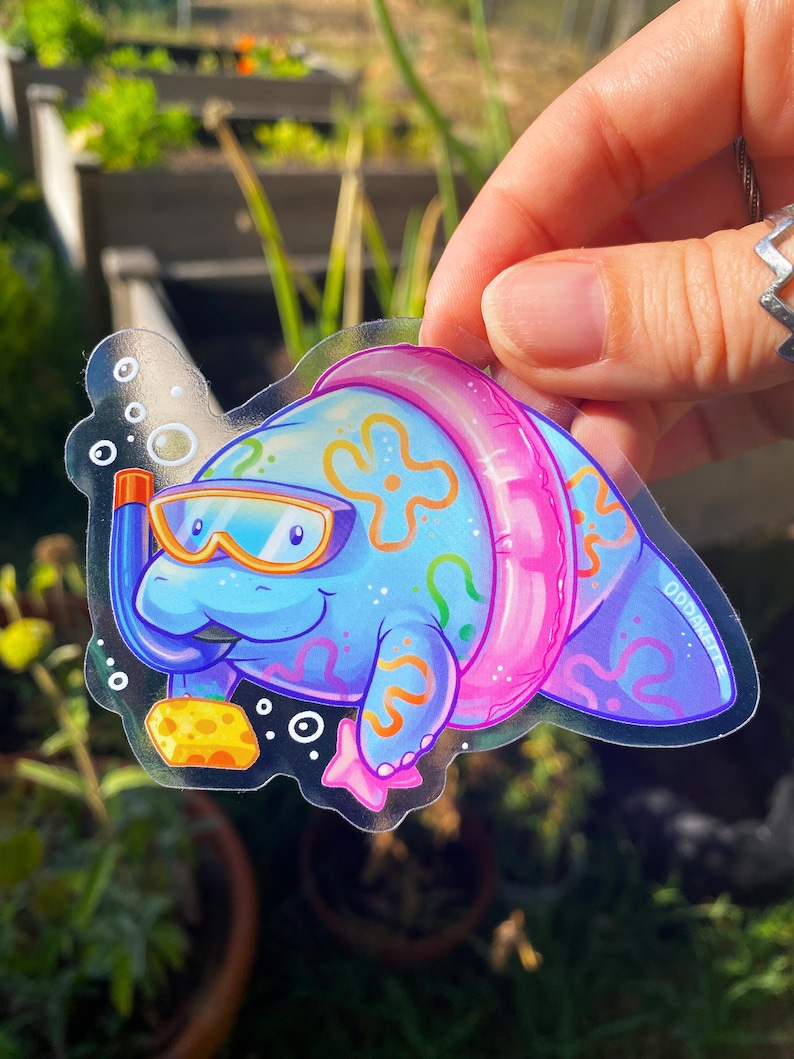May include: A clear sticker featuring a cartoon manatee wearing a pink inflatable ring and goggles. The manatee is blue with a yellow sponge and pink starfish. The sticker has the text "Oddskate" in the bottom right corner.