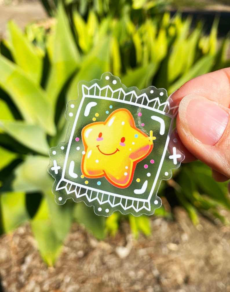 Puede incluir: Una pegatina transparente con una estrella amarilla con una cara sonriente y confeti de colores dentro de un marco blanco. La pegatina tiene un borde blanco festoneado.