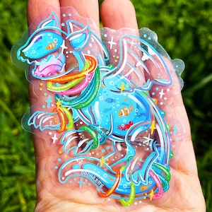 May include: A blue and white cartoon dragon with rainbow rings around its body. The dragon is surrounded by small fish and a red crab. The sticker is on a clear background and has the text "Oddareite" on the bottom.