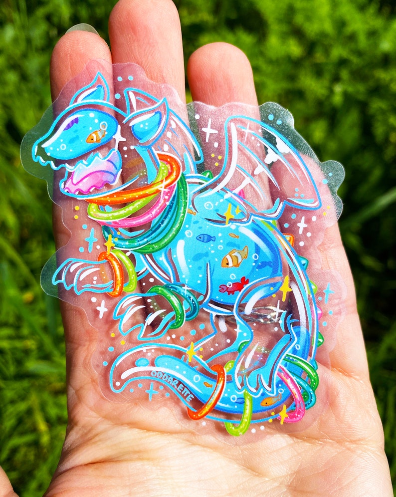 May include: A blue and white cartoon dragon with rainbow rings around its body. The dragon is surrounded by small fish and a red crab. The sticker is on a clear background and has the text "Oddareite" on the bottom.