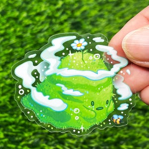 May include: A clear sticker featuring a cartoon green planet with a smiling face, a small white flower, and a bee. The planet is surrounded by white clouds and sparkles.