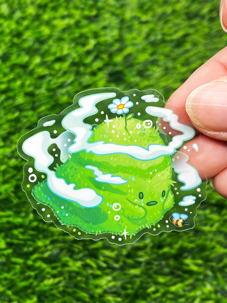May include: A clear sticker featuring a cartoon green planet with a smiling face, a small white flower, and a bee. The planet is surrounded by white clouds and sparkles.