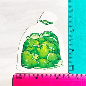 May include: A sticker featuring a clear bag filled with cartoon green frogs. The frogs have various poses, some with their tongues out. The sticker is approximately 3 inches tall and 2 inches wide.