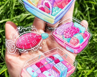 Può includere: Tre adesivi in ​​plastica trasparente con illustrazioni di cibo giapponese. Gli adesivi mostrano rotoli di sushi rosa e blu, un pesce rosa e una scatola bento rosa e blu con un cuore rosa e il testo "Oddaretie".