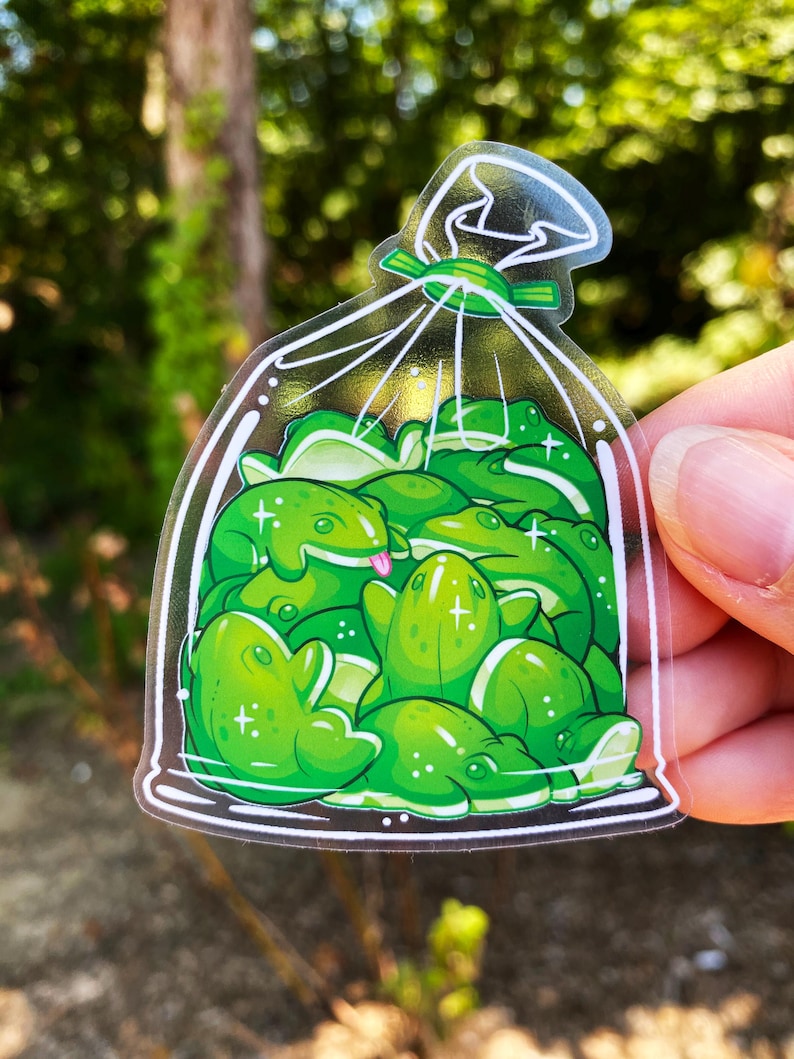 May include: A clear sticker featuring a bag of cartoon frogs. The frogs are various shades of green with white highlights and a few have their tongues sticking out. The bag is tied with a green ribbon.