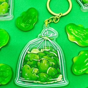 May include: A green frog-themed keychain with a bag of frogs design. The keychain features a clear acrylic charm with a detailed illustration of multiple green frog figures inside a transparent bag, with a green keyring.