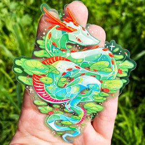 May include: A green and red dragon sticker with a translucent background. The dragon is curled up with its head resting on its tail. The dragon is surrounded by green lily pads and red ladybugs.