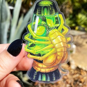 May include: A clear plastic sticker of a green lava lamp with a yellow alien creature inside. The alien has a segmented body and long, thin arms.