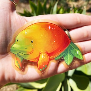 K&ouml;nnte beinhalten: Ein transparenter Aufkleber mit einem Cartoon-Manati in einem gr&uuml;n-orangen Farbverlauf. Der Manati l&auml;chelt und hat kleine, schwarze Augen. Der Aufkleber hat einen glitzernden, funkelnden Effekt und der Text "ODDARETTE" ist auf dem Aufkleber gedruckt.