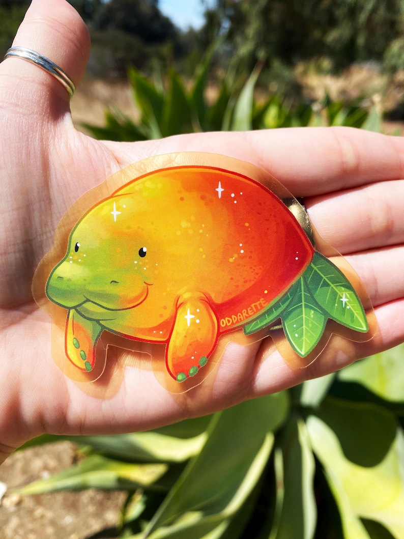 K&ouml;nnte beinhalten: Ein transparenter Aufkleber mit einem Cartoon-Manati in einem gr&uuml;n-orangen Farbverlauf. Der Manati l&auml;chelt und hat kleine, schwarze Augen. Der Aufkleber hat einen glitzernden, funkelnden Effekt und der Text "ODDARETTE" ist auf dem Aufkleber gedruckt.