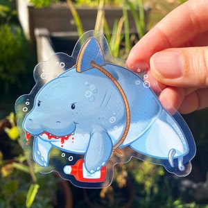 May include: A clear sticker with a cartoon illustration of a blue manatee with a red mouth and a bottle of hot sauce. The manatee is tied up with rope and has a hook in its tail.