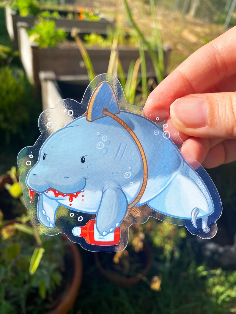 May include: A clear sticker with a cartoon illustration of a blue manatee with a red mouth and a bottle of hot sauce. The manatee is tied up with rope and has a hook in its tail.