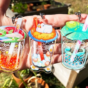 May include: Three clear acrylic bubble tea charms with different designs. The first charm features a fish and orange boba pearls. The second charm features a chicken in a nest with white and yellow boba pearls. The third charm features a frog on a lily pad with blue boba pearls.
