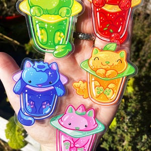 May include: Five colorful cat-shaped stickers with a clear background. Each sticker features a cat inside a clear cup with a different colored liquid and fruit. The cats are in various poses, including sitting, sleeping, and looking up.