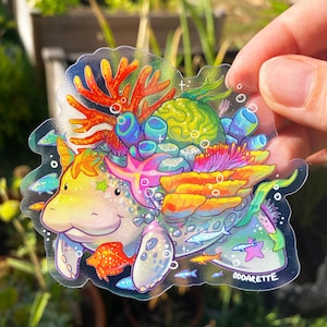 May include: A clear sticker featuring a colorful cartoon manatee with coral, seaweed, and fish. The manatee is surrounded by a rainbow of colors and has a sparkly texture. The sticker is signed "Oddarete".
