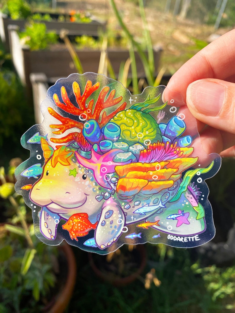May include: A clear sticker featuring a colorful cartoon manatee with coral, seaweed, and fish. The manatee is surrounded by a rainbow of colors and has a sparkly texture. The sticker is signed "Oddarete".